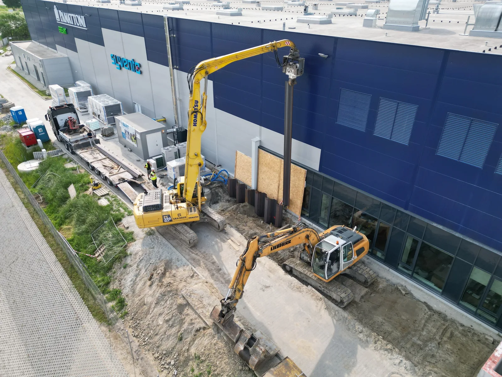 Koparka Komatsu z wibromłotem i pionową grodzicą przy obudowie wykopu obok hali.