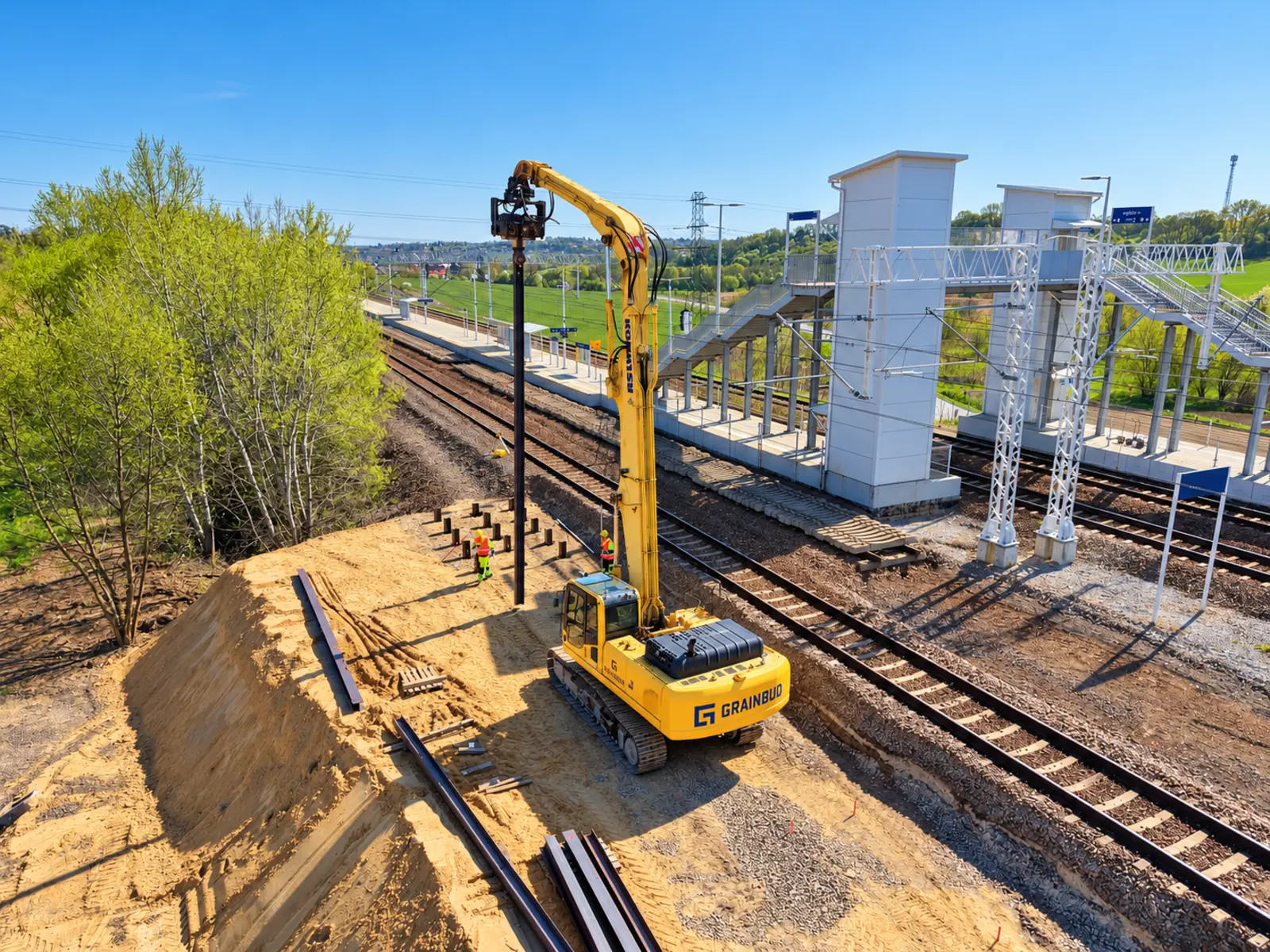 Komatsu z wibromłotem pogrąża profil stalowy przy posadowieniu pośrednim schodów peronowych.