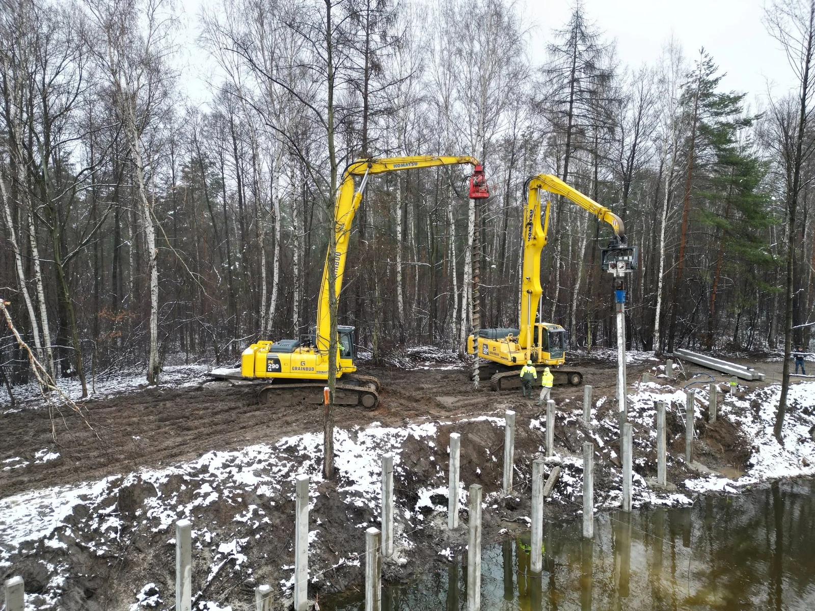 Dwie koparki Komatsu, jedna z wiertnicą, druga z wibromłotem, przy rzędzie pionowych elementów nad wodą.